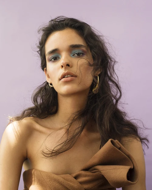 Portrait of a brunette female model with colorful eyeshadow, gold hoop earrings, and draped in a brown textile against a lilac background.
