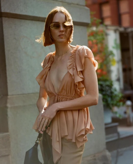 Woman in peach-colored ruffled blouse and sunglasses stands in an urban setting with buildings, greenery, and architecture visible in the background.