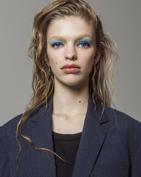 Young woman with wet blonde hair, blue eyeshadow, and a navy blazer in a studio portrait against a light gray background.