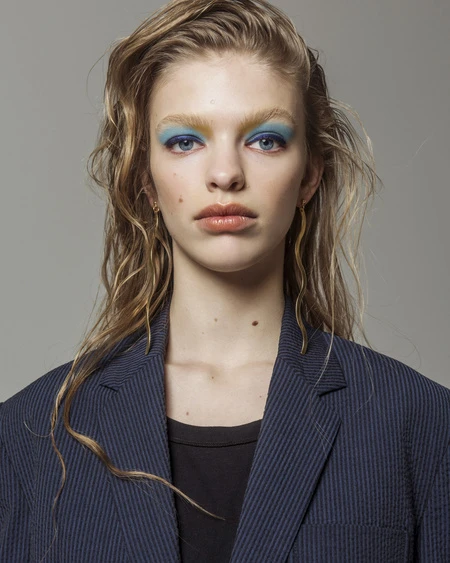 Young woman with wet blonde hair, blue eyeshadow, and a navy blazer in a studio portrait against a light gray background.