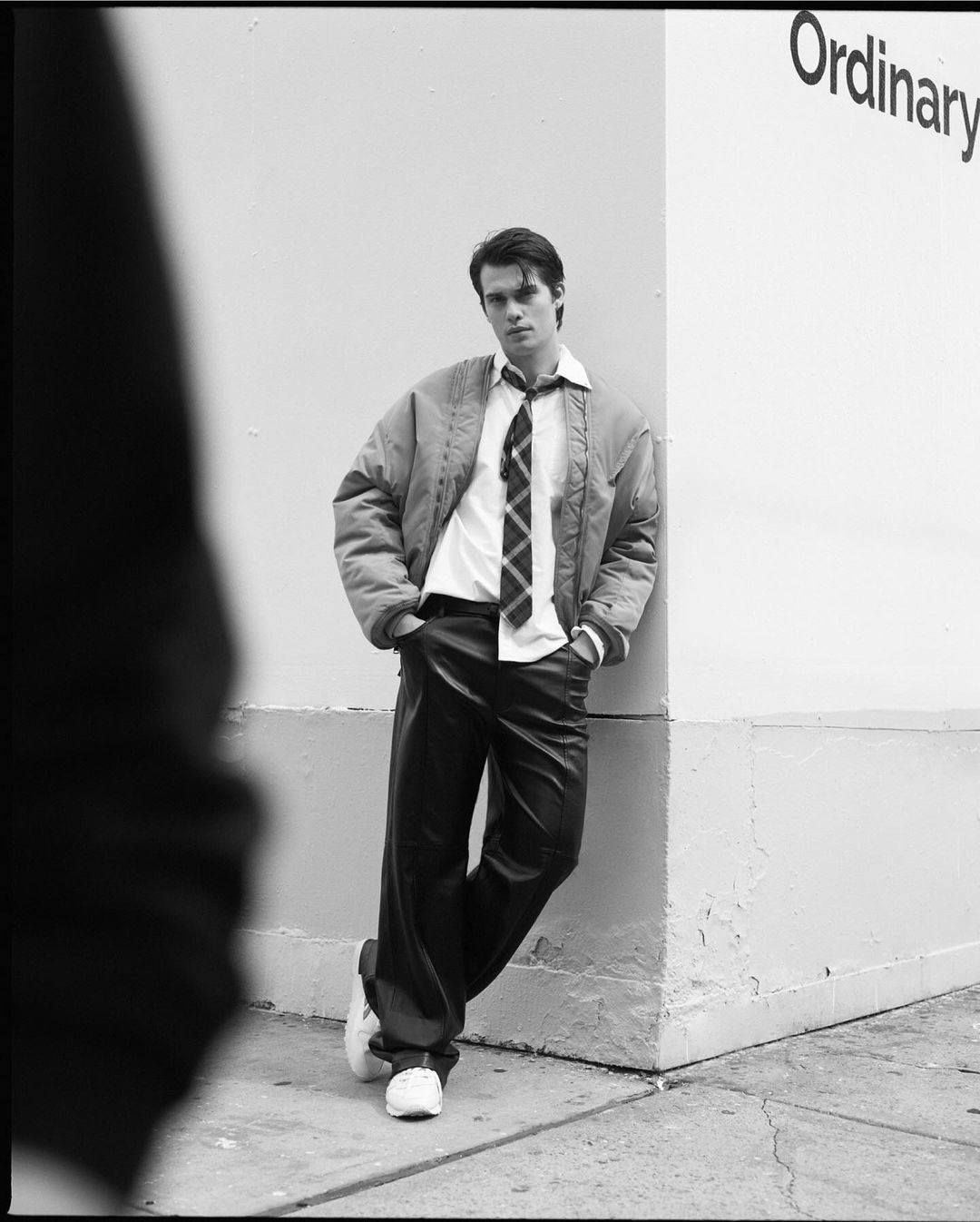 Monochrome portrait of a young man in an unzipped bomber jacket, white shirt, and tie, leaning against a weathered corner wall outdoors.