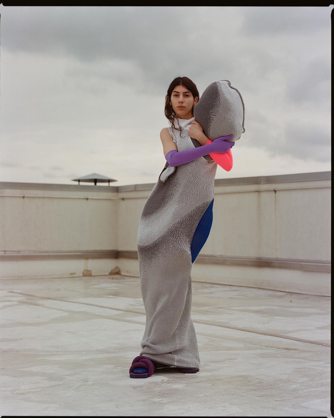 Woman in silver dress, purple gloves, and pink sleeve posing on a rooftop against a cloudy sky. Fashion editorial photograph.
