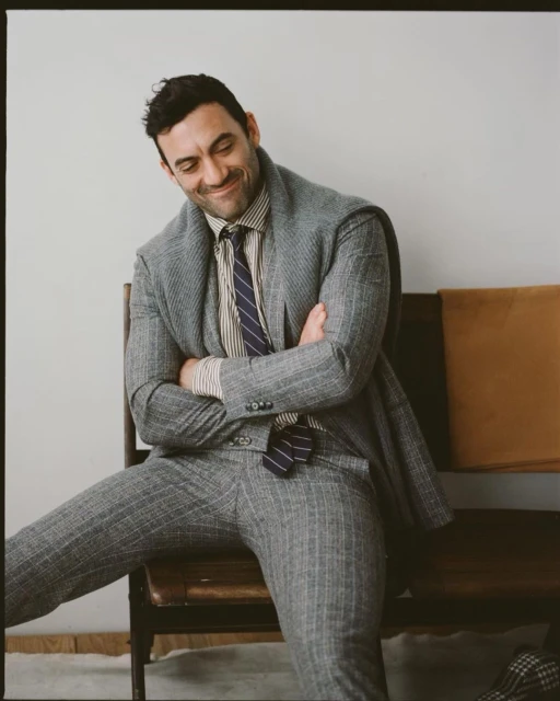 Adult man seated on a bench wearing a grey pinstripe suit, striped shirt, and striped tie in an indoor setting.