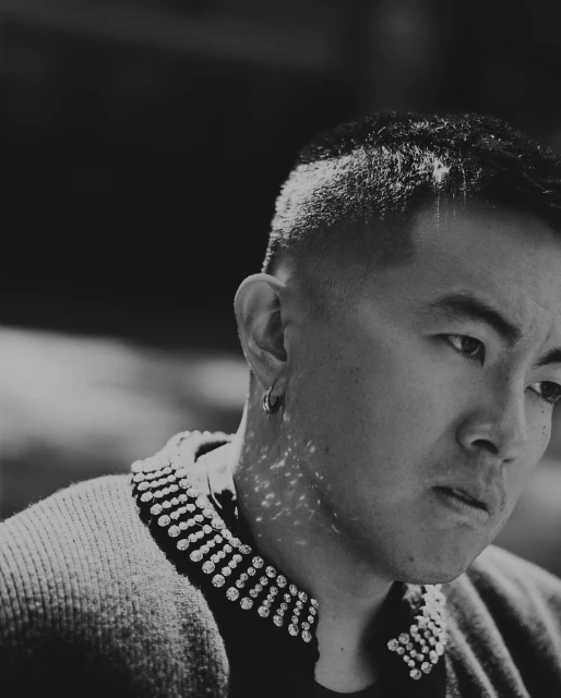 Monochrome portrait of an Asian man with short hair, wearing a sweater with jeweled embellishments around the collar.