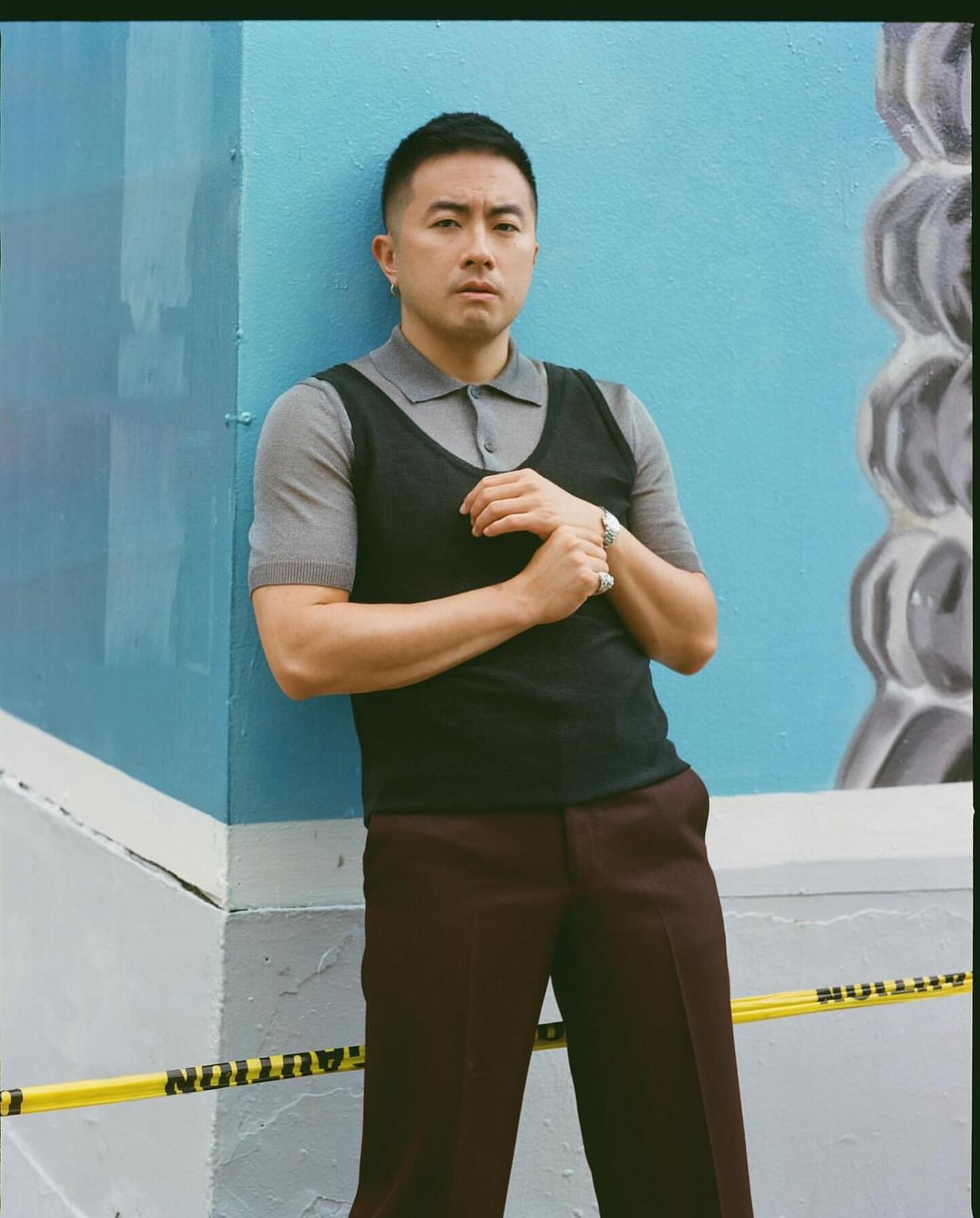 Stylish adult male in polo and vest leans against a vibrant blue wall with abstract patterns, separated by yellow caution tape.