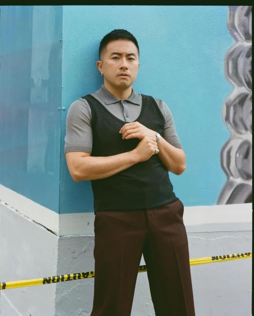 Stylish adult male in polo and vest leans against a vibrant blue wall with abstract patterns, separated by yellow caution tape.