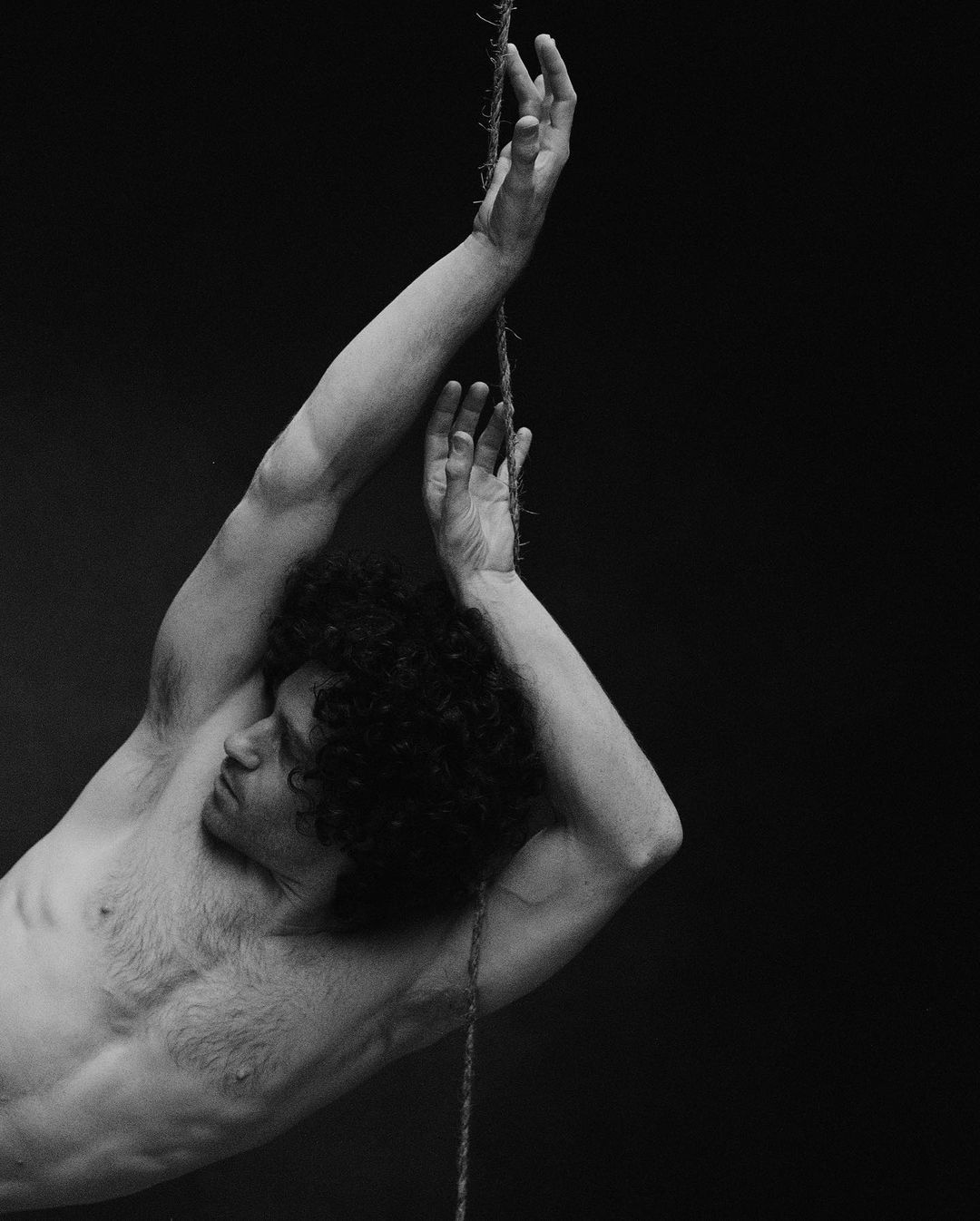 Striking black and white portrait of a shirtless adult male with curly hair, eyes closed, and hands reaching up to a rope.