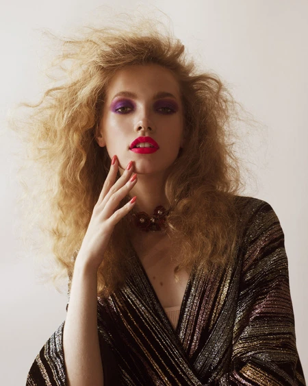 Close-up studio portrait of a fair-skinned young adult woman with curly blonde hair, wearing colorful makeup and a striped top.