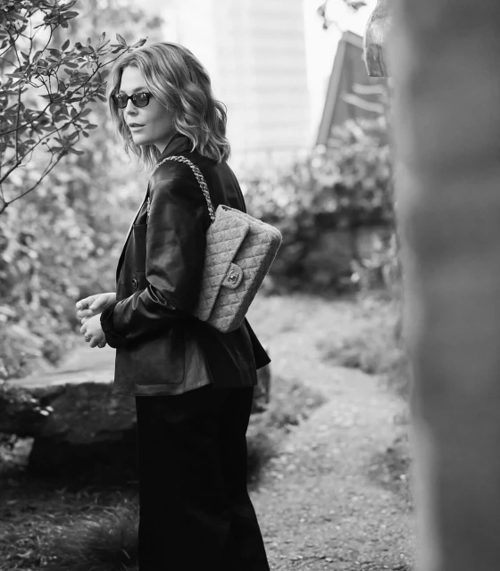 Adult woman with short wavy hair wearing sunglasses, jacket, and skirt, walking outdoors carrying a Chanel quilted shoulder bag.