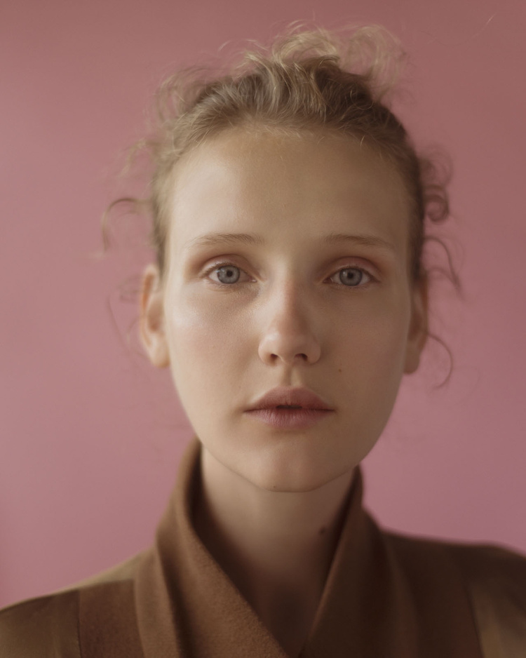 Portrait of a young woman with fair skin and light hair, wearing a brown jacket, against a pink background in a studio setting.