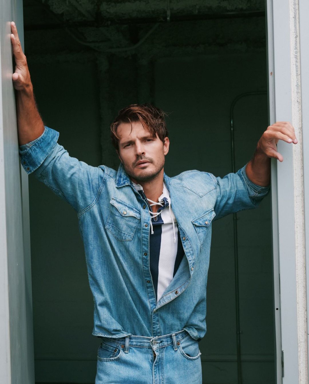 Man wearing a denim shirt and matching jeans leaning against a doorway, photographed by Matthew Priestley