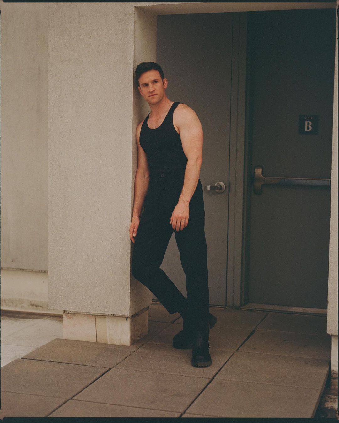 Man wearing a black tank top and trousers leaning against a doorway in a portrait by Matthew Priestley