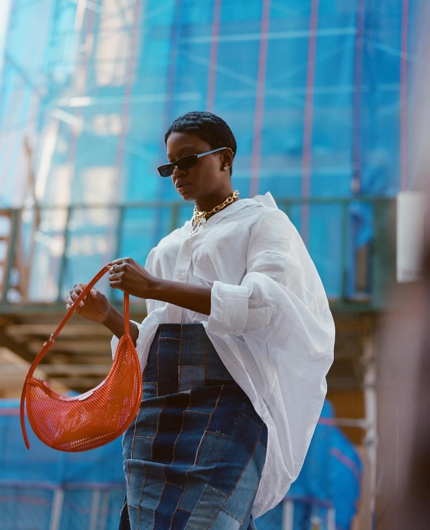 Stylish woman in white shirt and patchwork denim skirt poses outdoors holding red purse against a blue construction background.