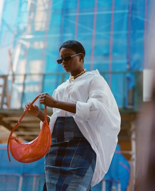 Stylish woman in white shirt and patchwork denim skirt poses outdoors holding red purse against a blue construction background.