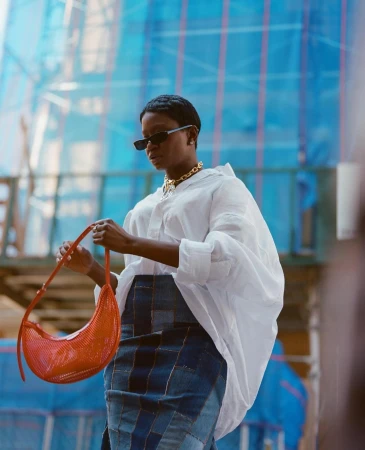 Stylish woman in white shirt and patchwork denim skirt poses outdoors holding red purse against a blue construction background.