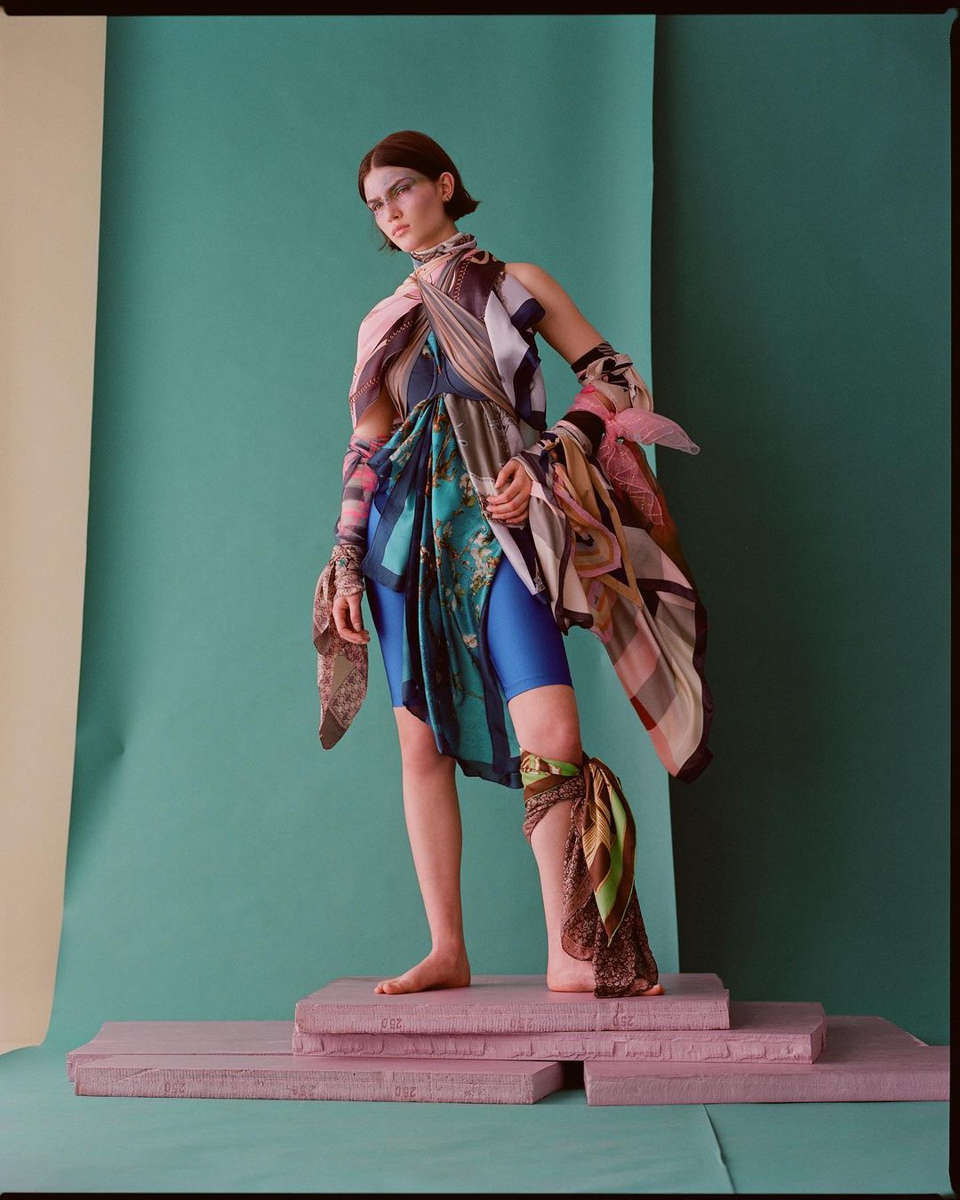 A young woman stands on pink platforms in a studio wearing bike shorts and draped in patterned scarves against a teal backdrop.