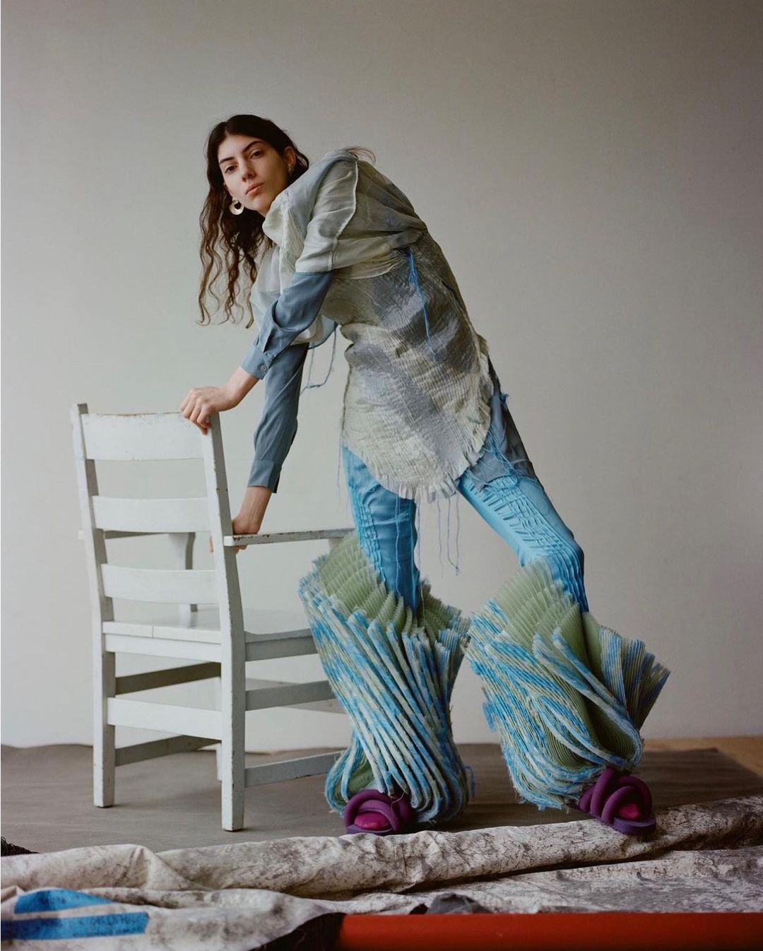 Woman strikes a pose leaning on a chair in blue jeans, an open-weave top, and voluminous shoes with purple soles in a studio setting.