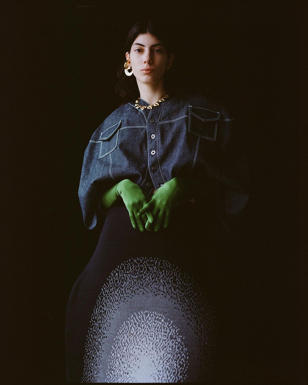 Fashion portrait of a young woman with gold jewelry wearing a denim jacket with green gloves in front of a dark background.