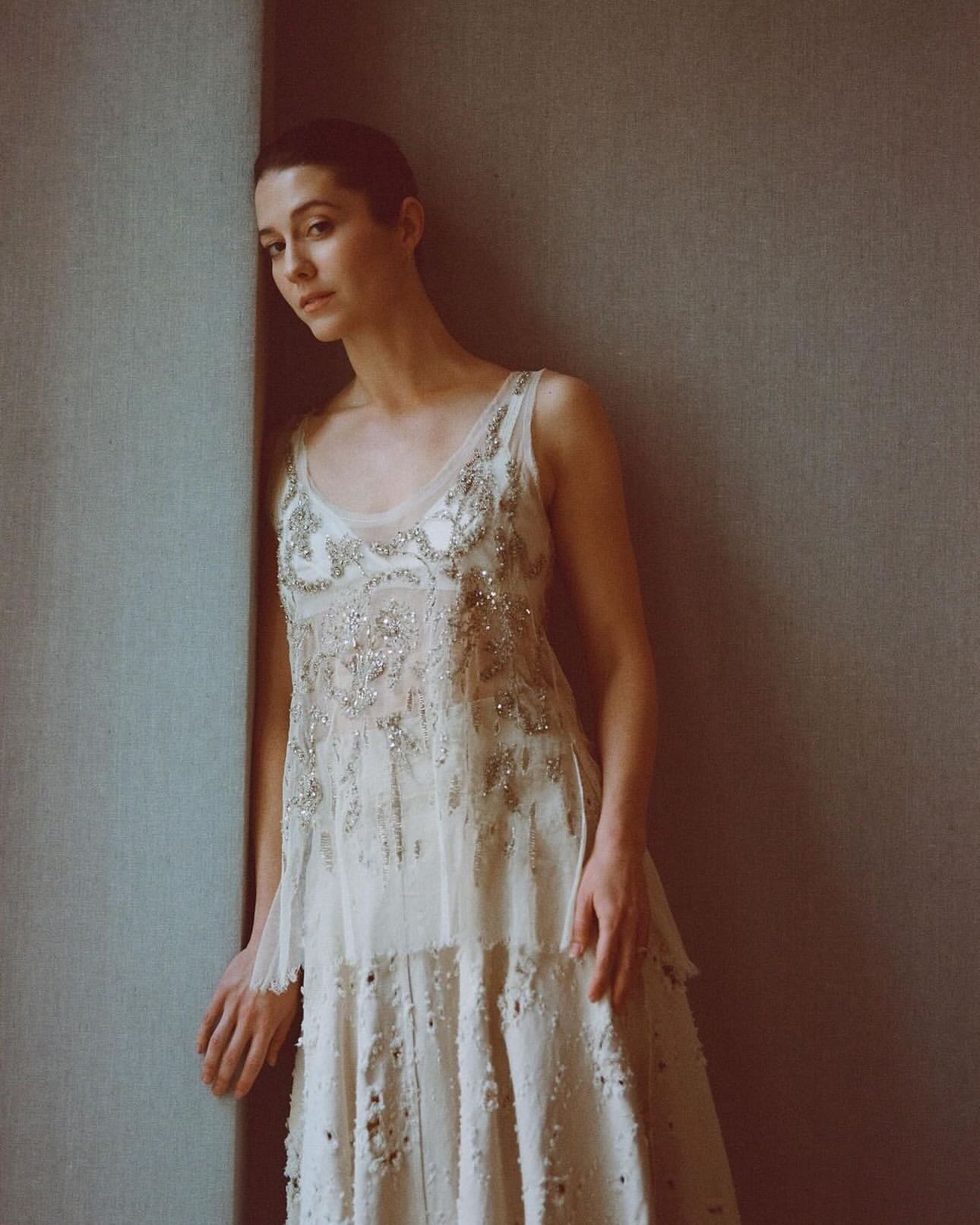 Portrait of a young adult woman in an ornate cream-colored dress leaning against a gray wall in a studio setting, looking at camera.
