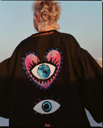 Back view of a blonde adult female wearing a black jacket with a heart-shaped sequin eye design, photographed outdoors in natural lighting.