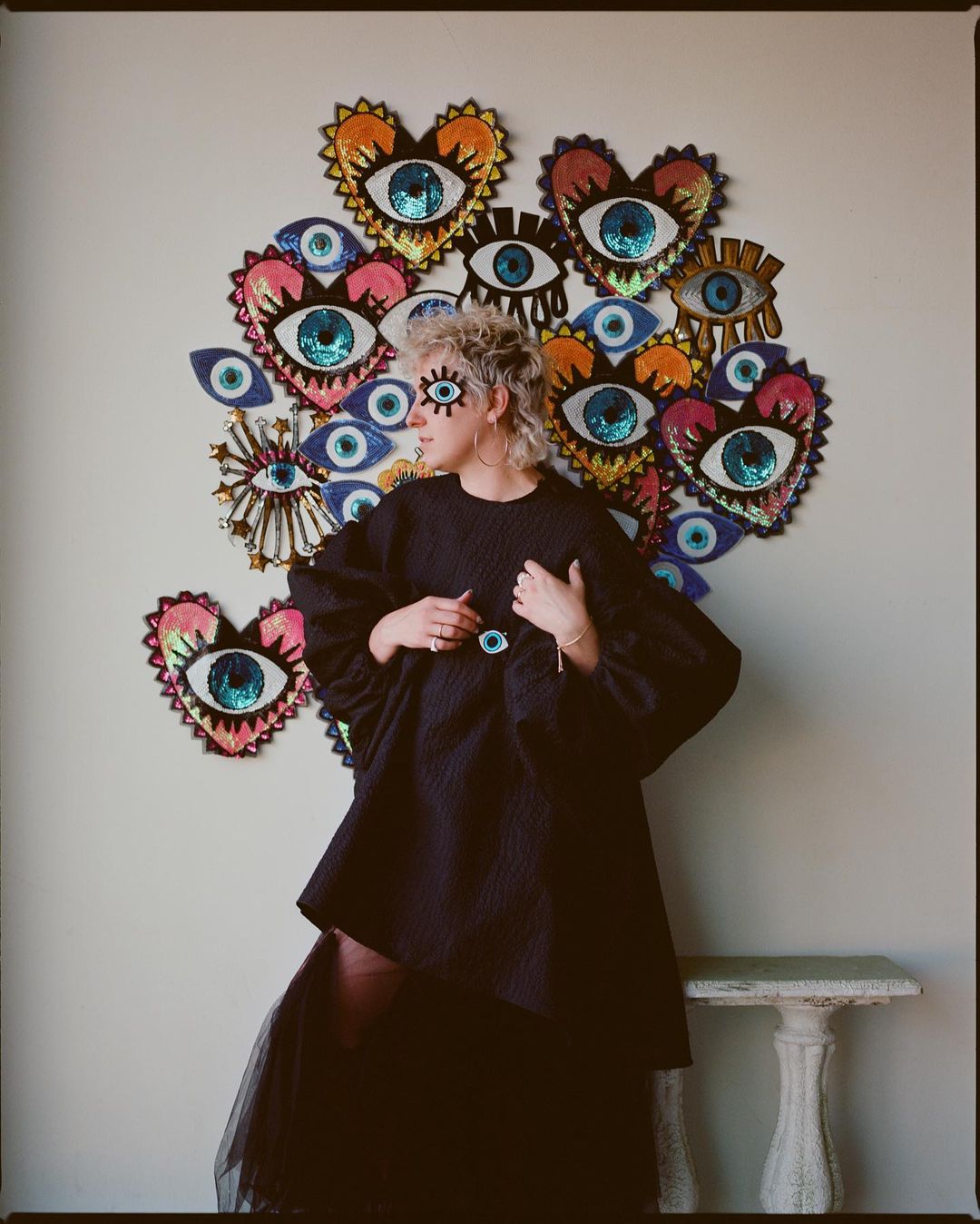 Eye-adorned woman wearing black outfit poses in front of wall with eye-themed art. Statement on fashion, beauty, and personal style.