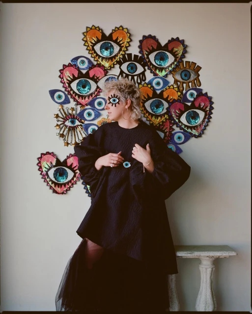 Eye-adorned woman wearing black outfit poses in front of wall with eye-themed art. Statement on fashion, beauty, and personal style.