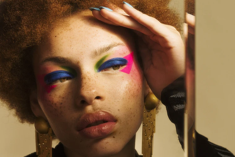 Close-up of a woman with auburn curly hair and blue eyeshadow, accented with pink and green makeup, wearing large gold earrings.