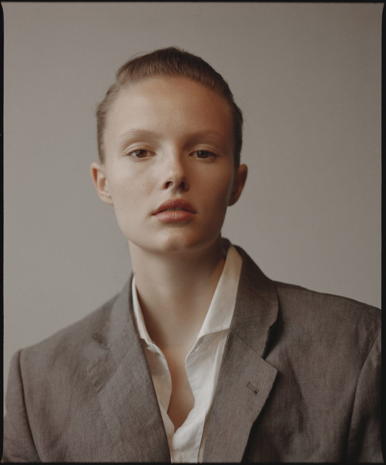 Portrait of a woman with slicked-back hair wearing a grey blazer and white shirt against a neutral background.