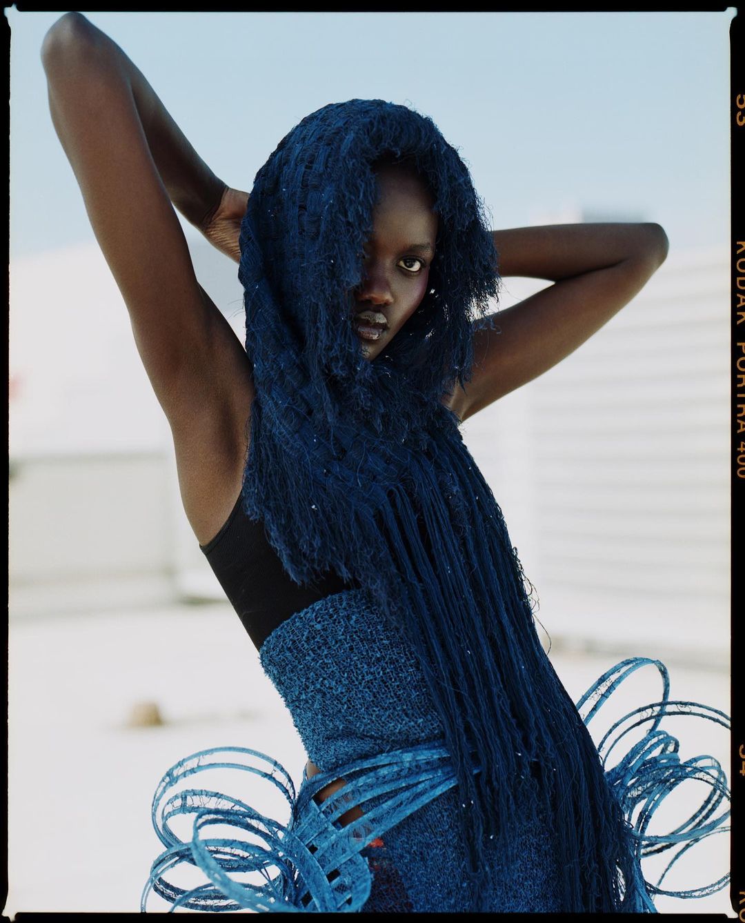 Stunning studio portrait of a dark-skinned woman in a blue, textured dress with fringed detailing and her arms raised overhead.