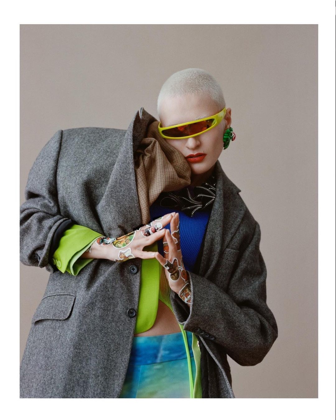 Fashionable woman with a shaved head wearing futuristic yellow sunglasses, a grey jacket, and statement earrings in a studio setting.
