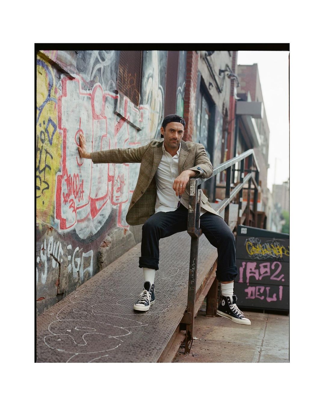 A man in a blazer and cap poses on a metal ramp with graffiti wall, wearing high-top sneakers, showcasing an urban fashion style.