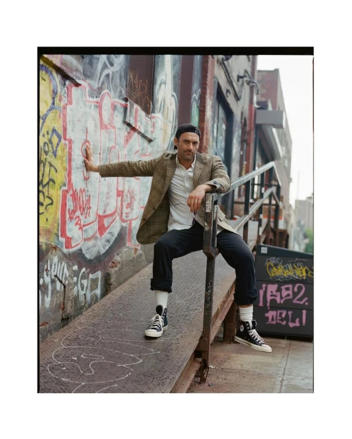 A man in a blazer and cap poses on a metal ramp with graffiti wall, wearing high-top sneakers, showcasing an urban fashion style.
