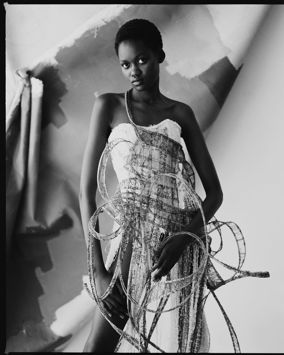 Monochrome portrait of a Black woman in a white dress adorned with film strips against an abstract painted backdrop in a photo studio.