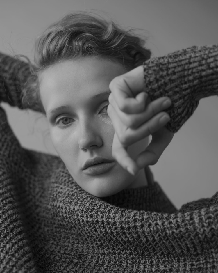 Monochrome portrait of a woman in a knitted sweater, gazing directly at the camera with her hands near her face.