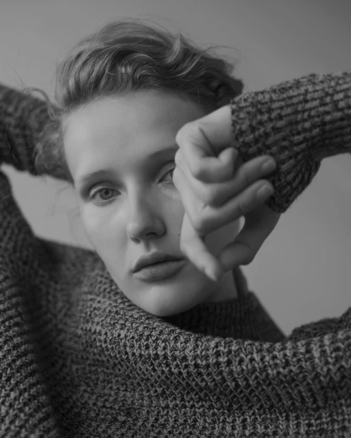 Monochrome portrait of a woman in a knitted sweater, gazing directly at the camera with her hands near her face.