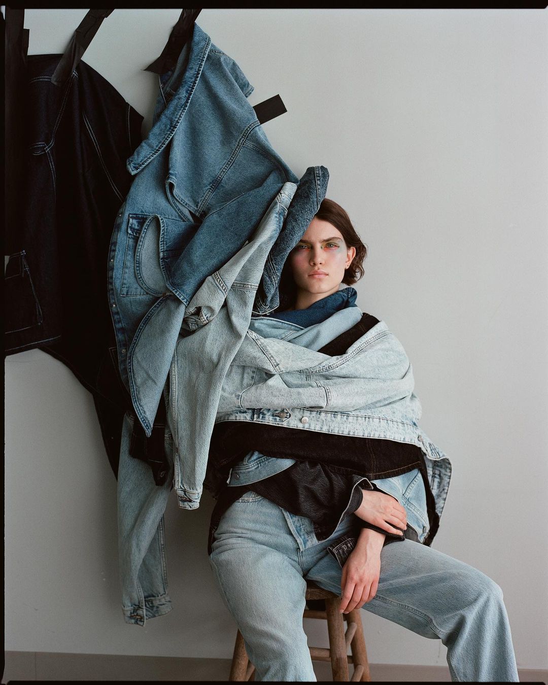 Young woman in denim outfit sits on stool, draped in denim jackets and jeans, gazing at camera against a neutral grey backdrop.