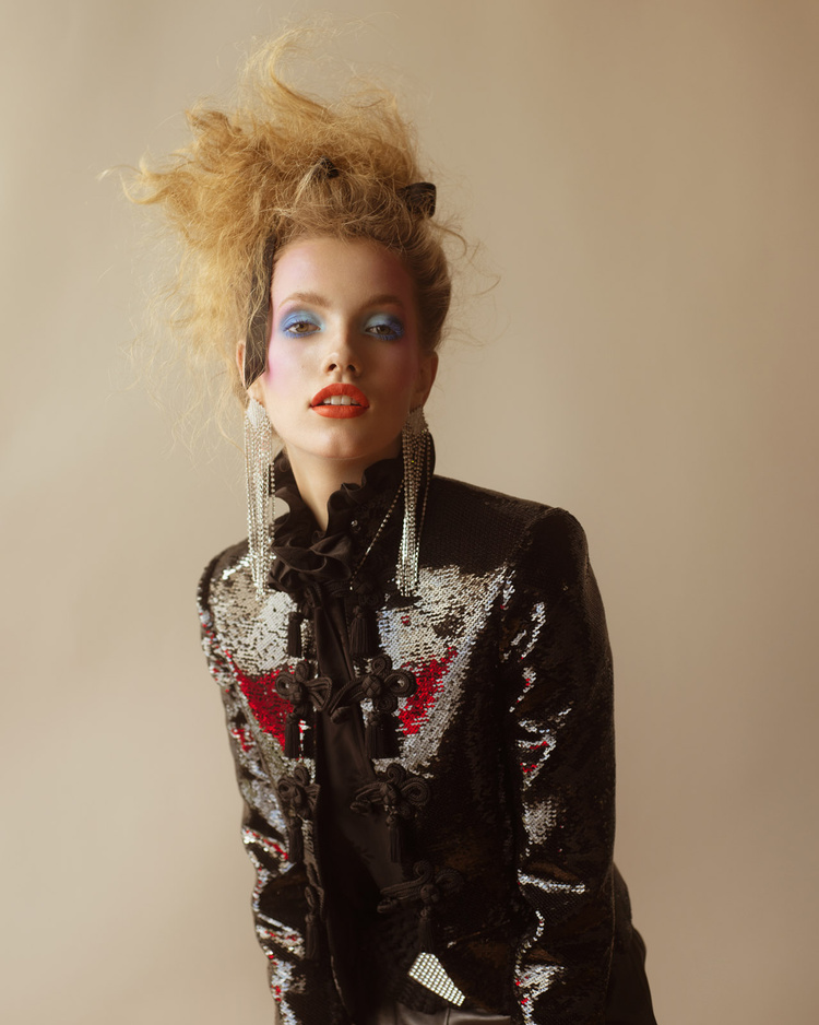 Eye-level portrait of a blonde adult female wearing a black sequin jacket, dramatic makeup, and long silver earrings against a tan background.