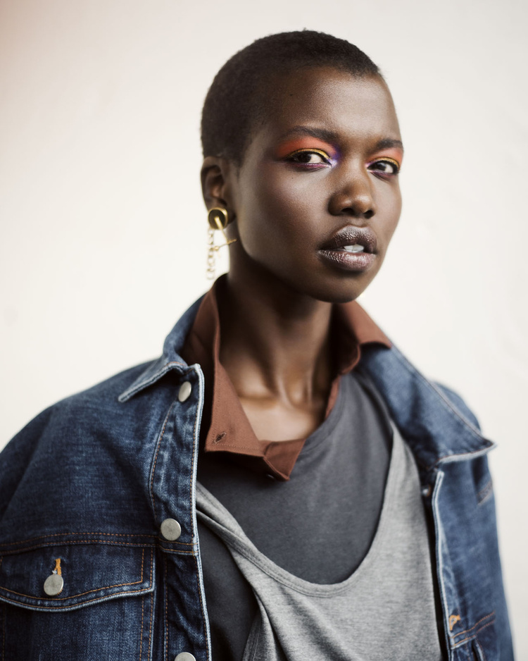 Portrait of a Black woman with short hair wearing a denim jacket and colorful eye makeup against a white backdrop.