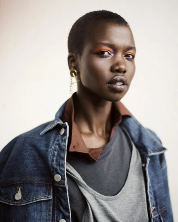 Portrait of a Black woman with short hair wearing a denim jacket and colorful eye makeup against a white backdrop.