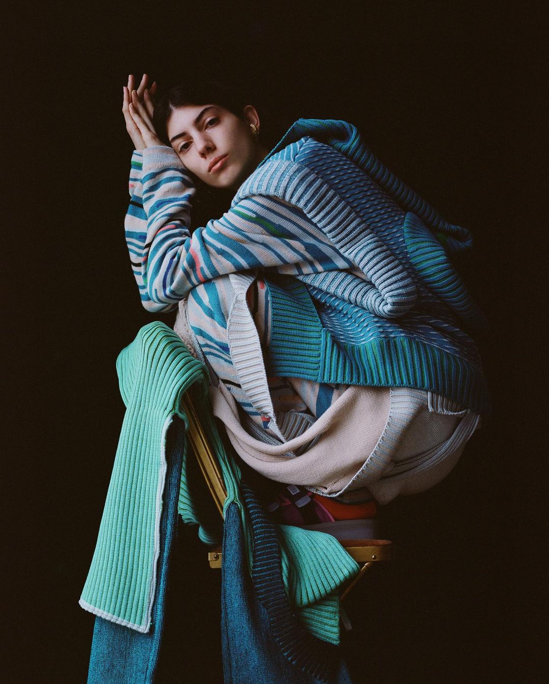 An adult female poses seated in a studio, wearing a colorful knit sweater and skirt, with a neutral expression against a dark background.