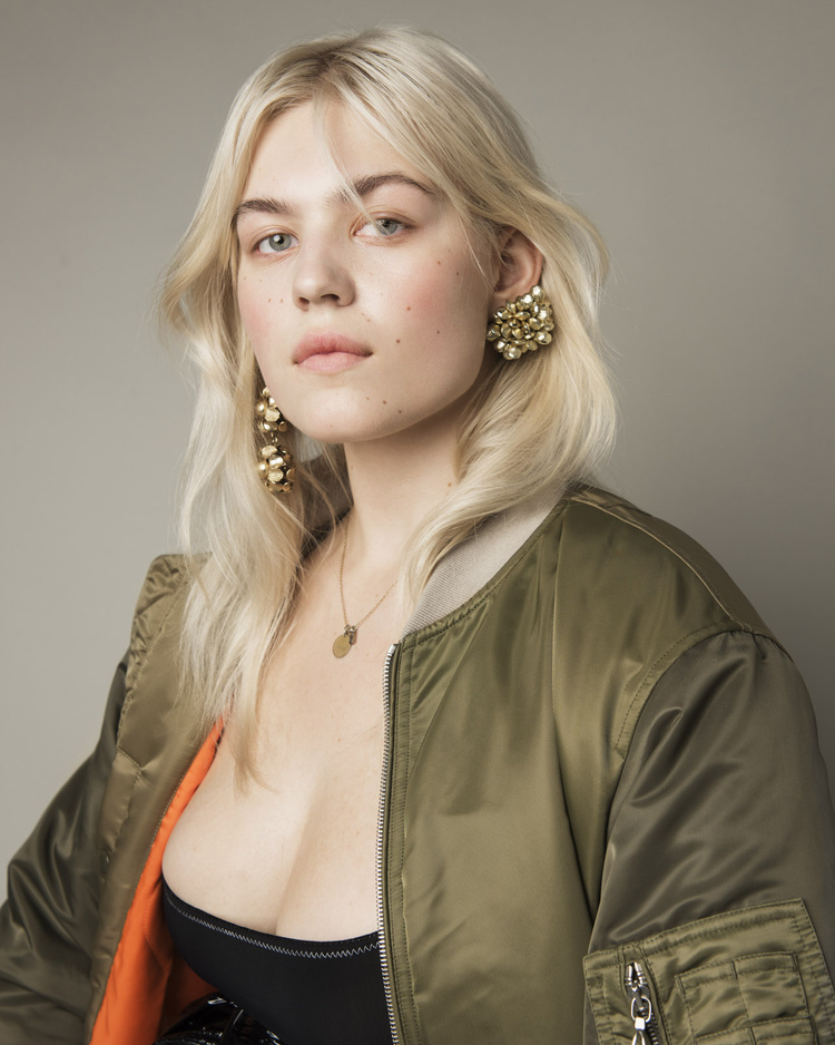 Portrait of a young woman with blonde hair wearing a green bomber jacket, earrings, and a necklace against a grey background.