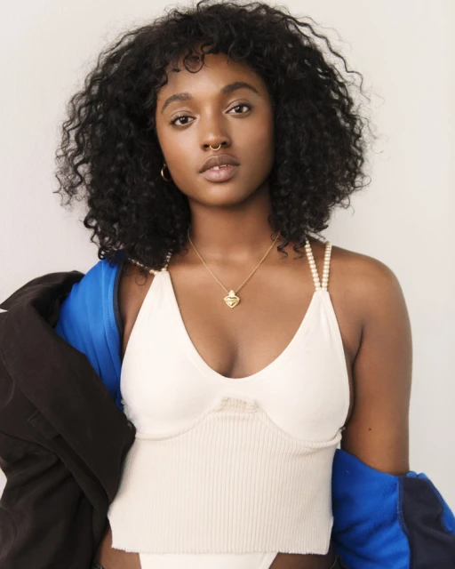 Close-up of a stylish young Black woman with curly hair and a septum piercing, wearing a white top and gold necklace in a studio setting.