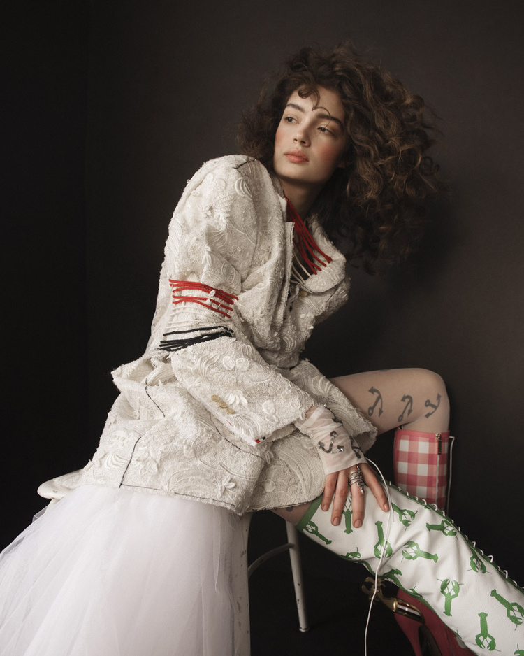 A curly-haired model in white textured jacket with red trim poses in a studio setting with playful green lobster-printed boots.