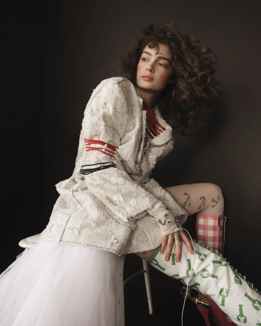 A curly-haired model in white textured jacket with red trim poses in a studio setting with playful green lobster-printed boots.