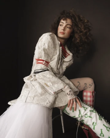 A curly-haired model in white textured jacket with red trim poses in a studio setting with playful green lobster-printed boots.