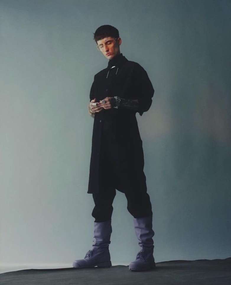 Portrait of a young adult male in a black outfit and boots, standing indoors against a muted gray backdrop in a fashion style.