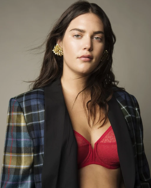 Brunette woman in a plaid jacket and red bra posing in front of a gray backdrop. She is wearing gold earrings.