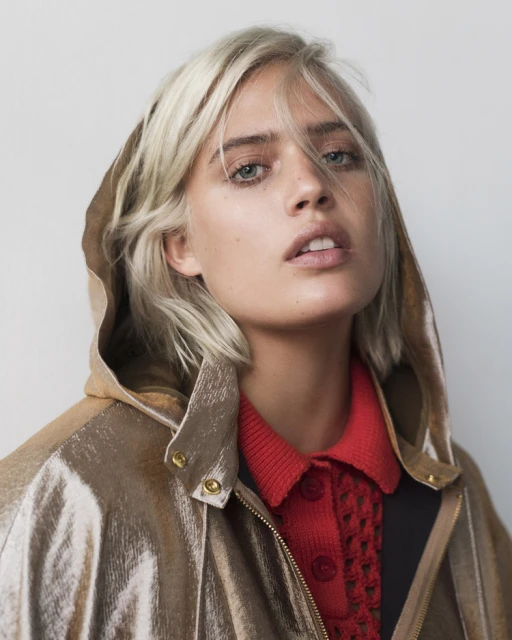 Close-up portrait of a blond-haired woman wearing a metallic gold hooded jacket and red top, posing against a neutral backdrop.