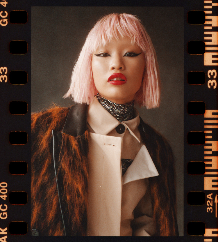 Portrait of an asian woman with pink hair, red lipstick, and a tan coat in front of a dark gray background. Film border is visible.