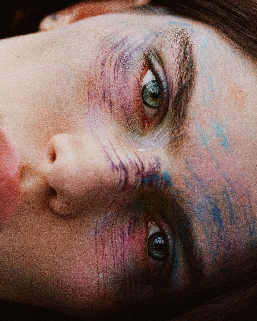 Extreme close-up of an adult female's face with colorful abstract brushstroke makeup. Focused eyes, light skin tone, and portrait orientation.
